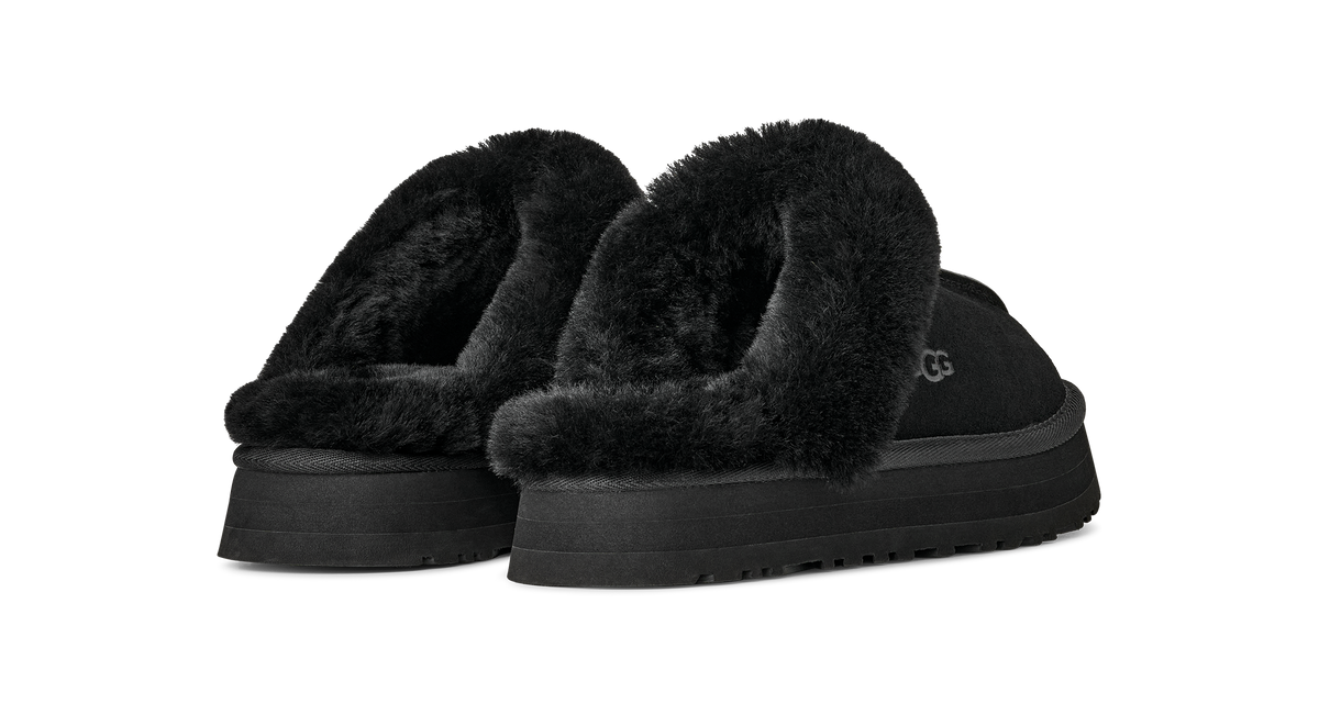 A pair of black UGG slip-on slippers with plush faux-fur trim and thick platform soles, shown against a solid white background.