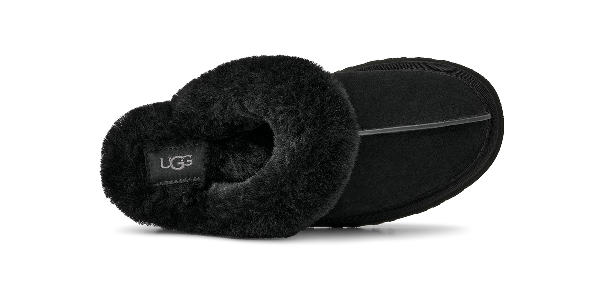 A black UGG slipper shown from a side profile view, featuring a soft black faux-shearling collar and a black suede-like body, with a small black UGG logo patch on the inside.