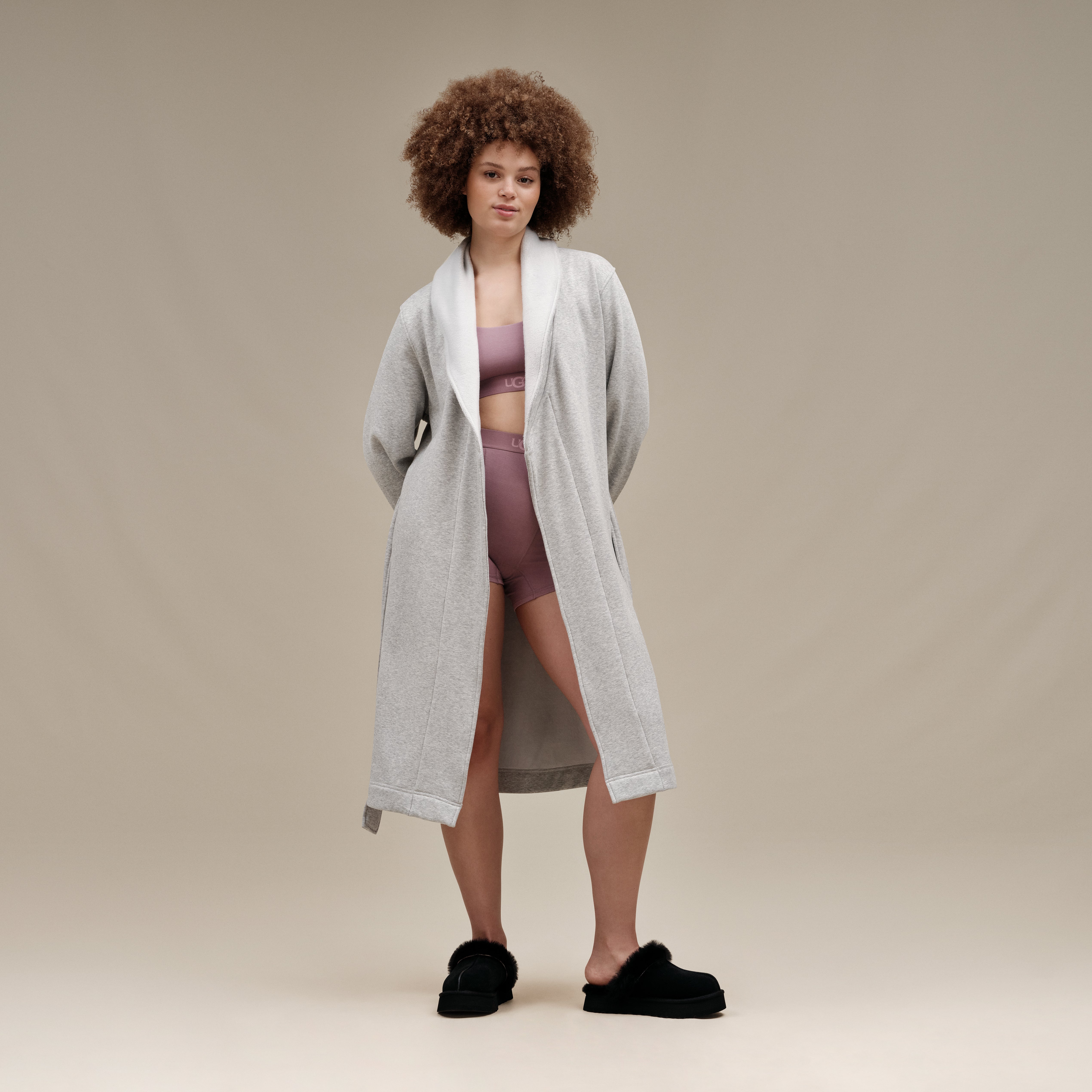 A woman stands against a plain beige studio background wearing a light gray open-front robe over a mauve-pink sports bra and matching high-waisted shorts, with black fuzzy slippers.