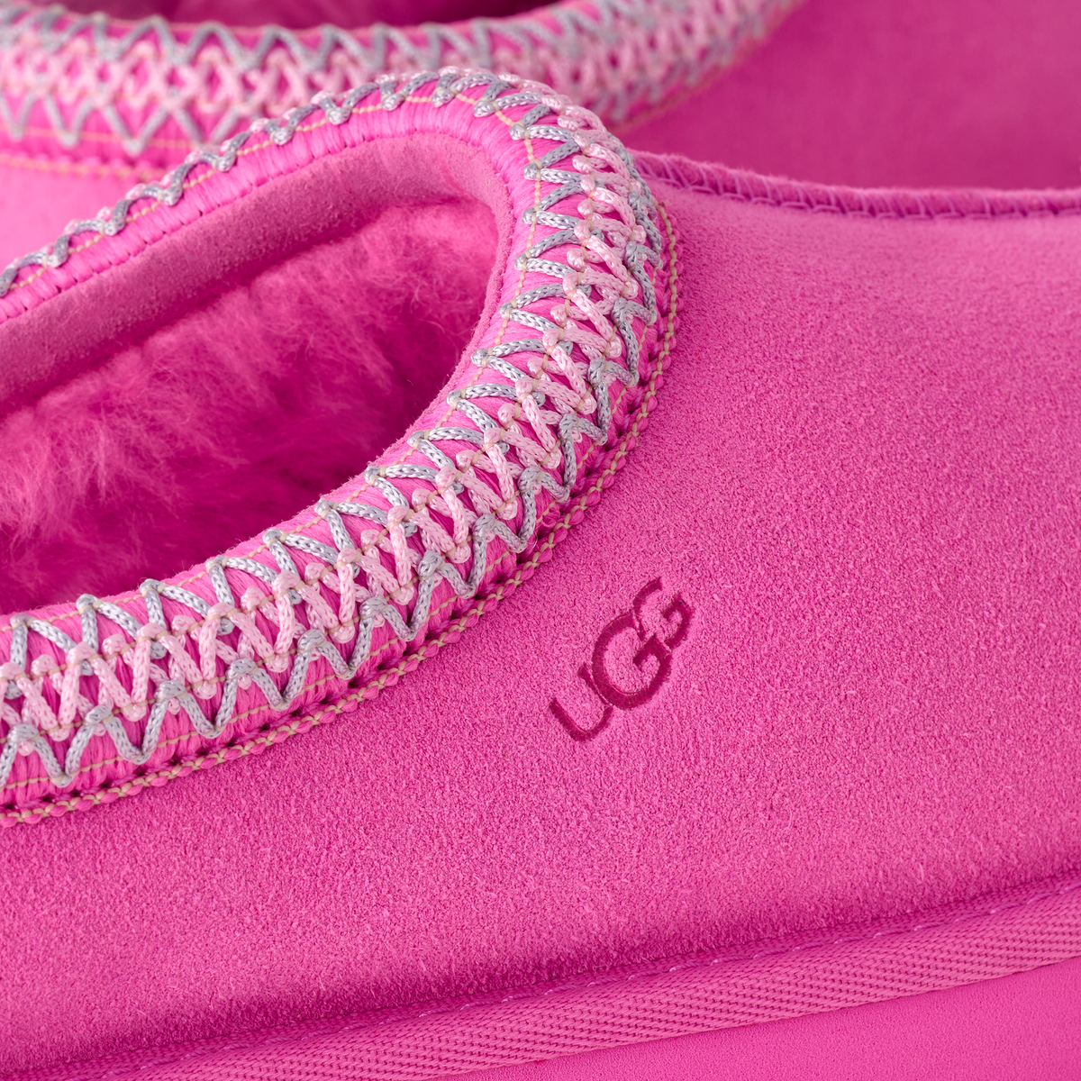 Close-up of a bright pink UGG slipper showing the suede-like exterior, fuzzy pink interior lining, and decorative zigzag trim around the opening.