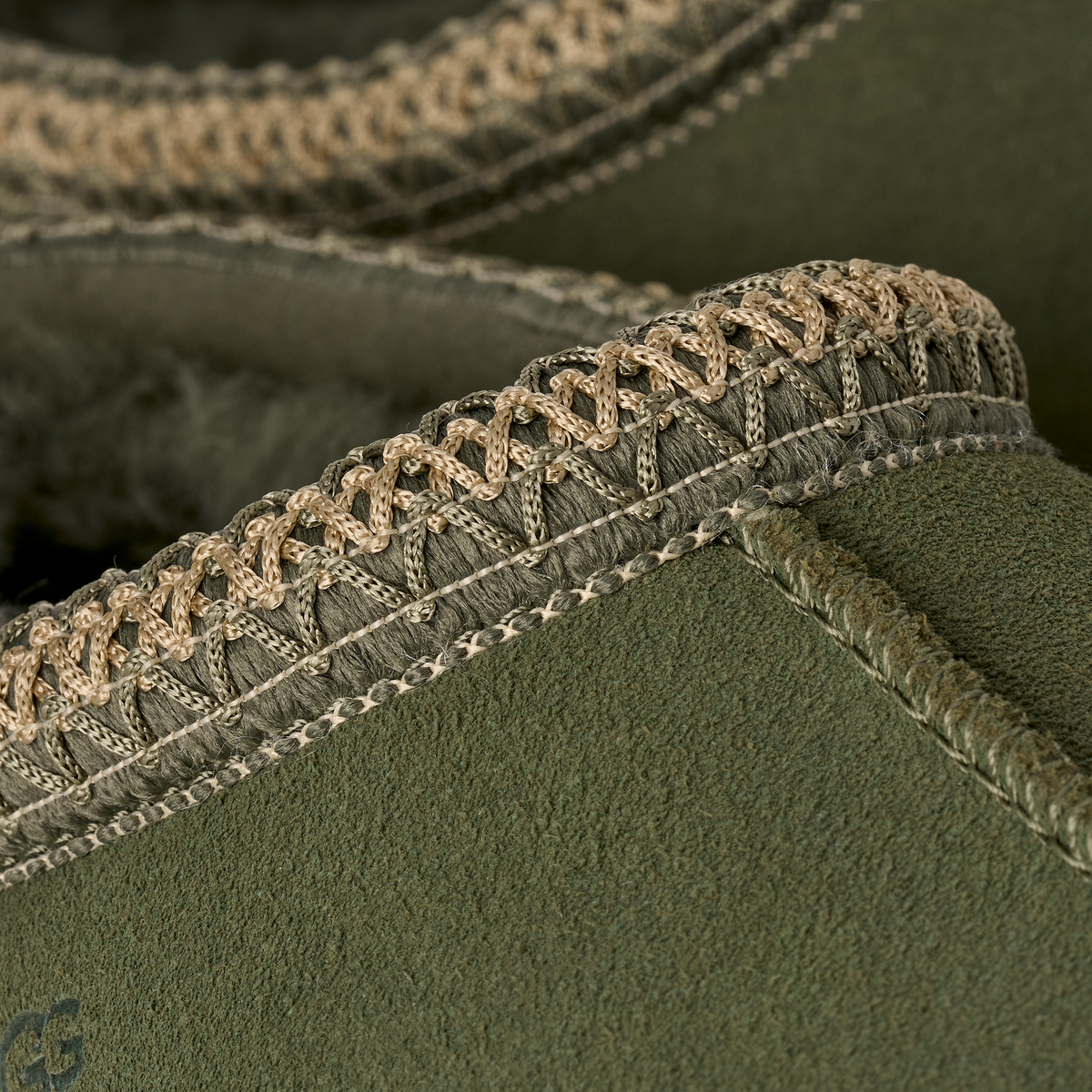 Close-up detail of an olive green suede slipper with intricate braided beige trim around the opening.