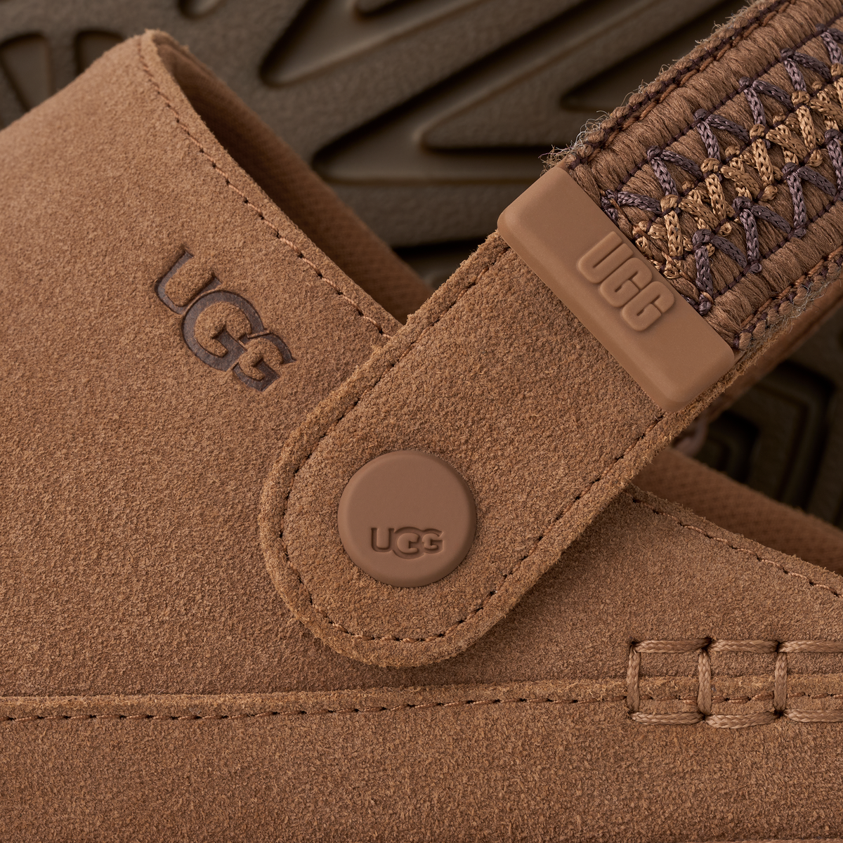 Close-up detail of a UGG boot in tan suede, showing the embossed UGG logo on the strap, a round UGG button, and a braided strap detail against a dark textured background.
