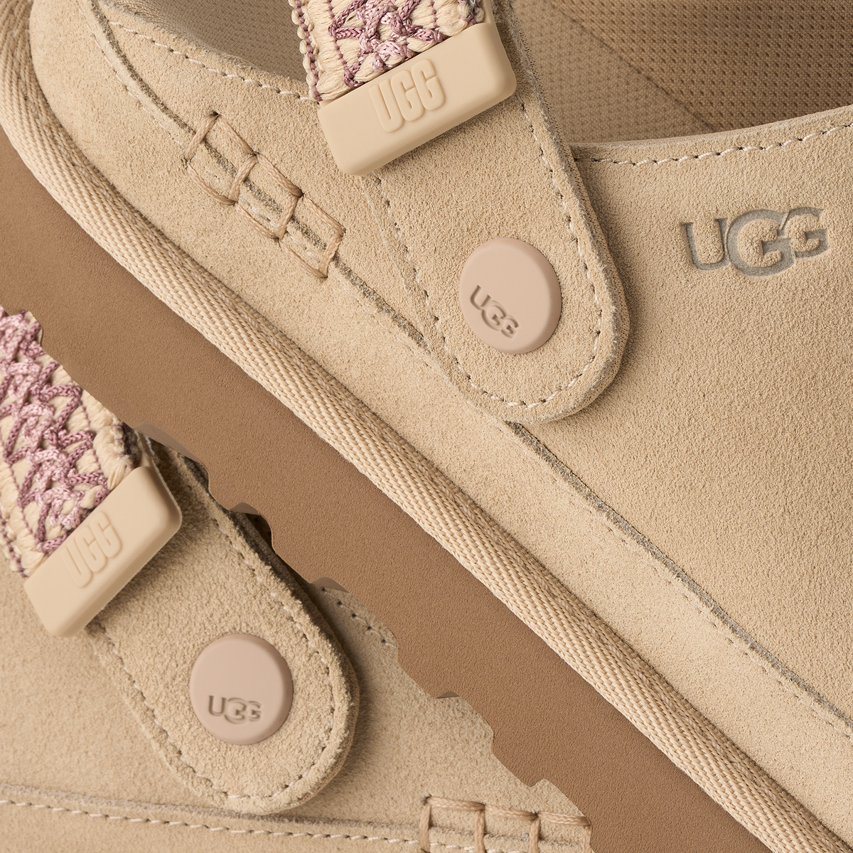 Close-up of a beige suede UGG shoe with matching beige strap closures, round UGG buttons, and patterned woven details on the straps.