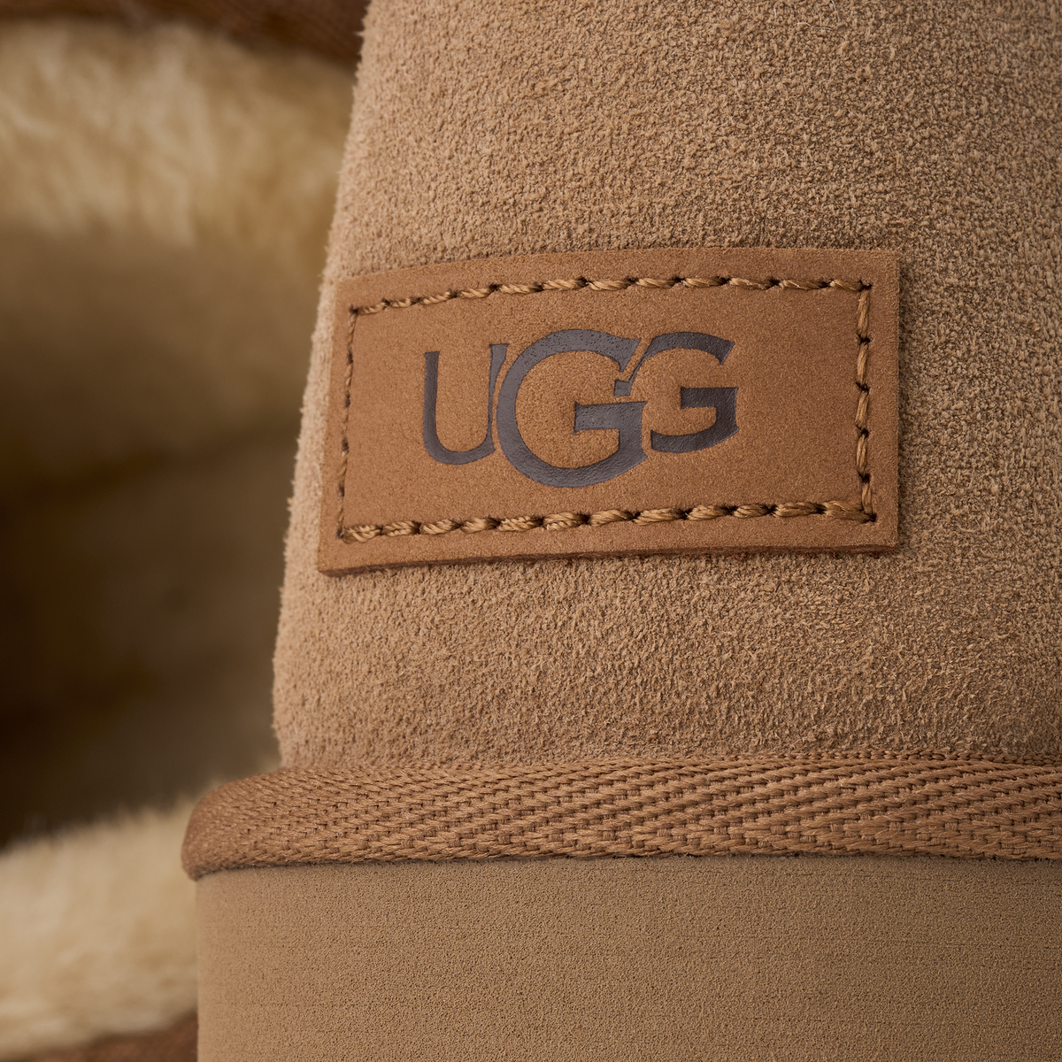 Close-up of a tan UGG boot showing the brand patch on the side, with suede-like texture, a braided band, and soft fur lining in the background.