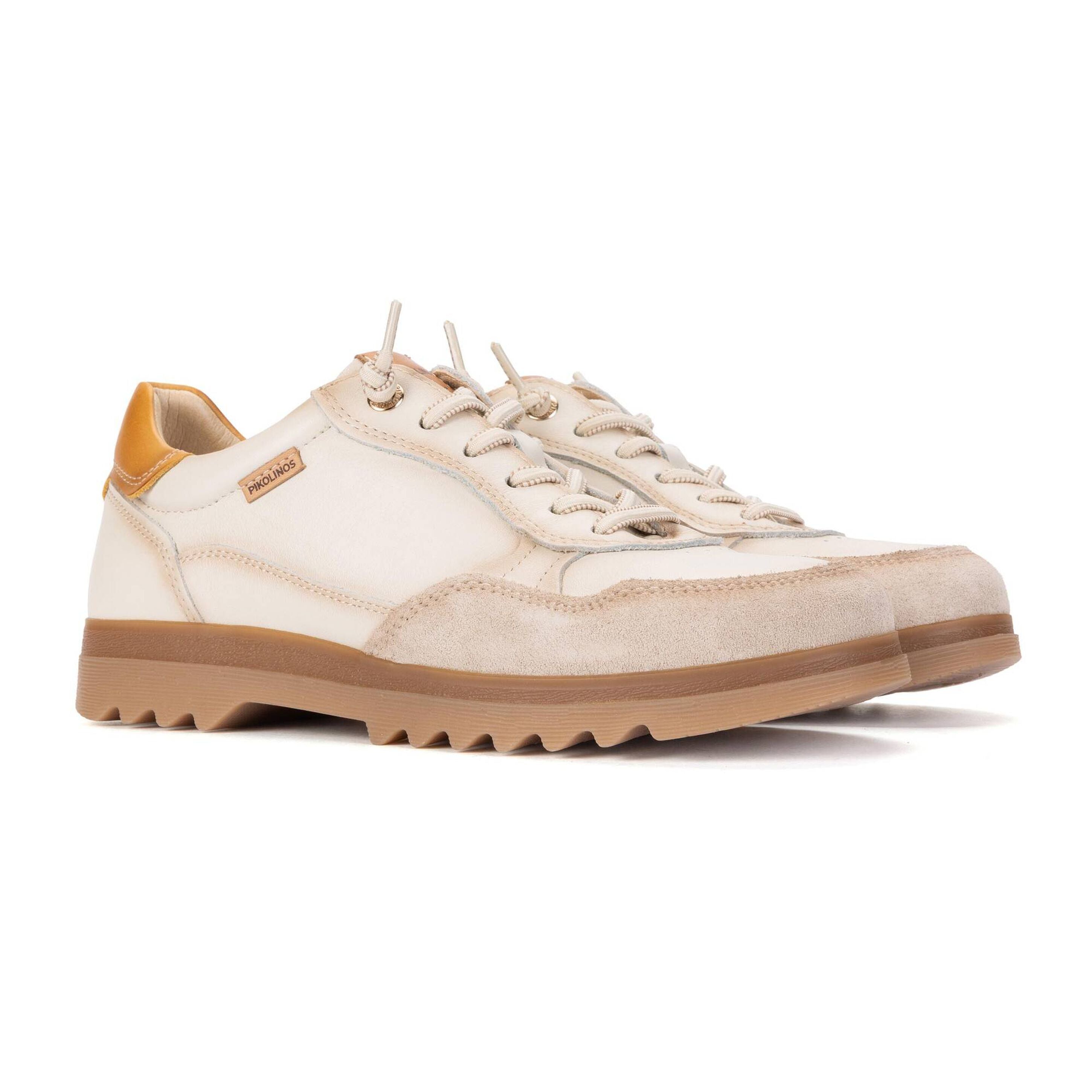 A low-top sneaker in off-white suede and canvas with beige laces, a tan sole, and a small brown leather brand tab on the side, photographed on a white background.