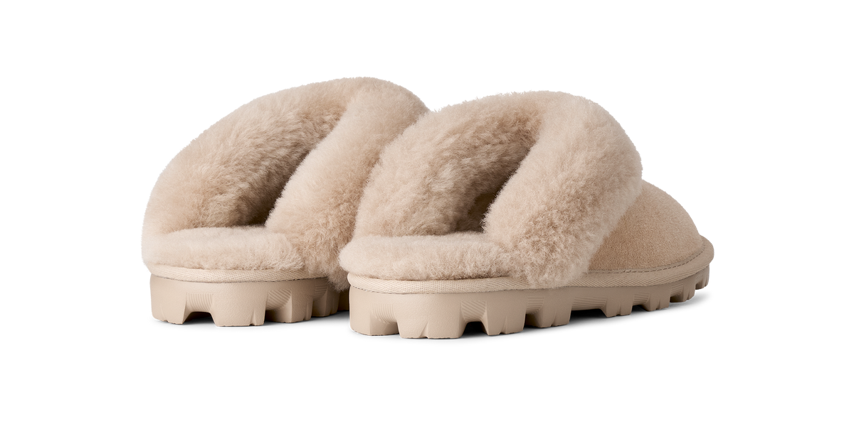 A pair of beige shearling-style slide slippers with thick, textured beige soles, shown from a three-quarters angle on a solid white background.