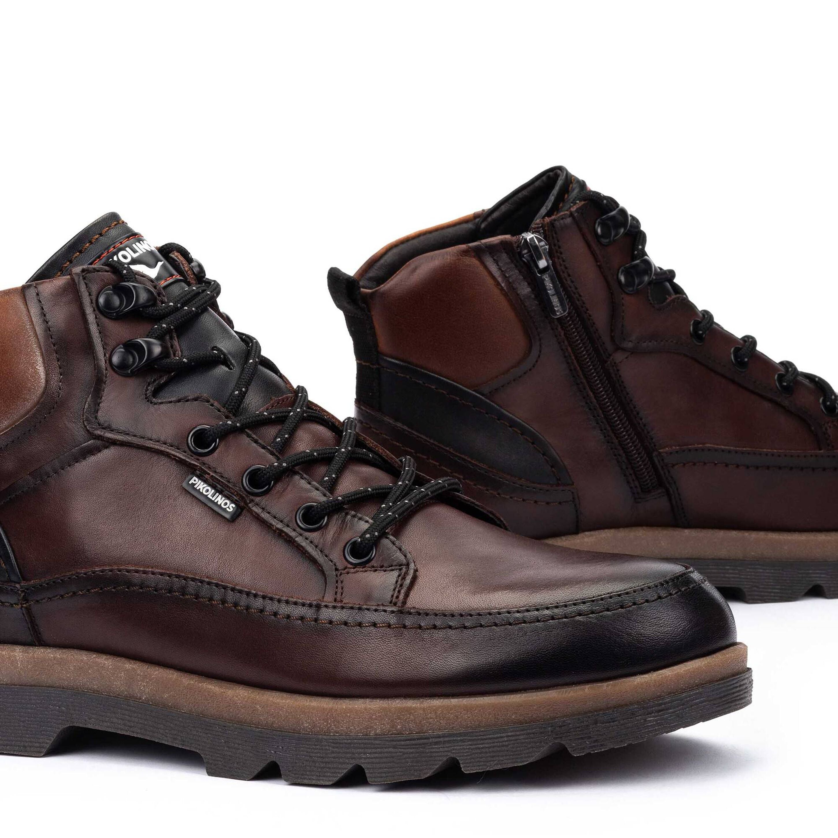 A pair of dark brown leather lace-up boots with black laces, black side panels, and a tan midsole, shown on a white background.