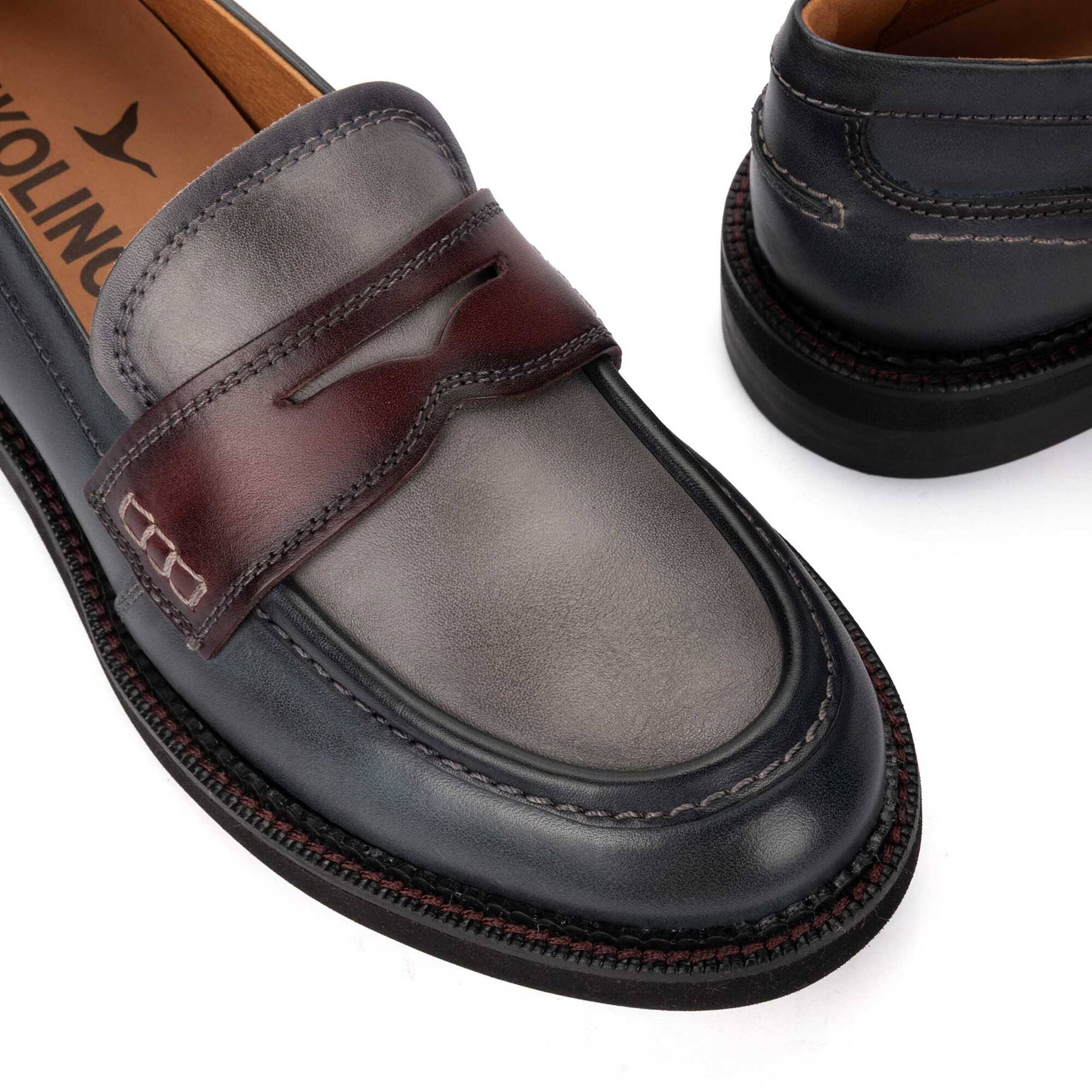 A close-up of a penny loafer in dark brown and black leather, showing the side profile with contrast stitching and a pull strap. A second shoe is partially visible in the background.