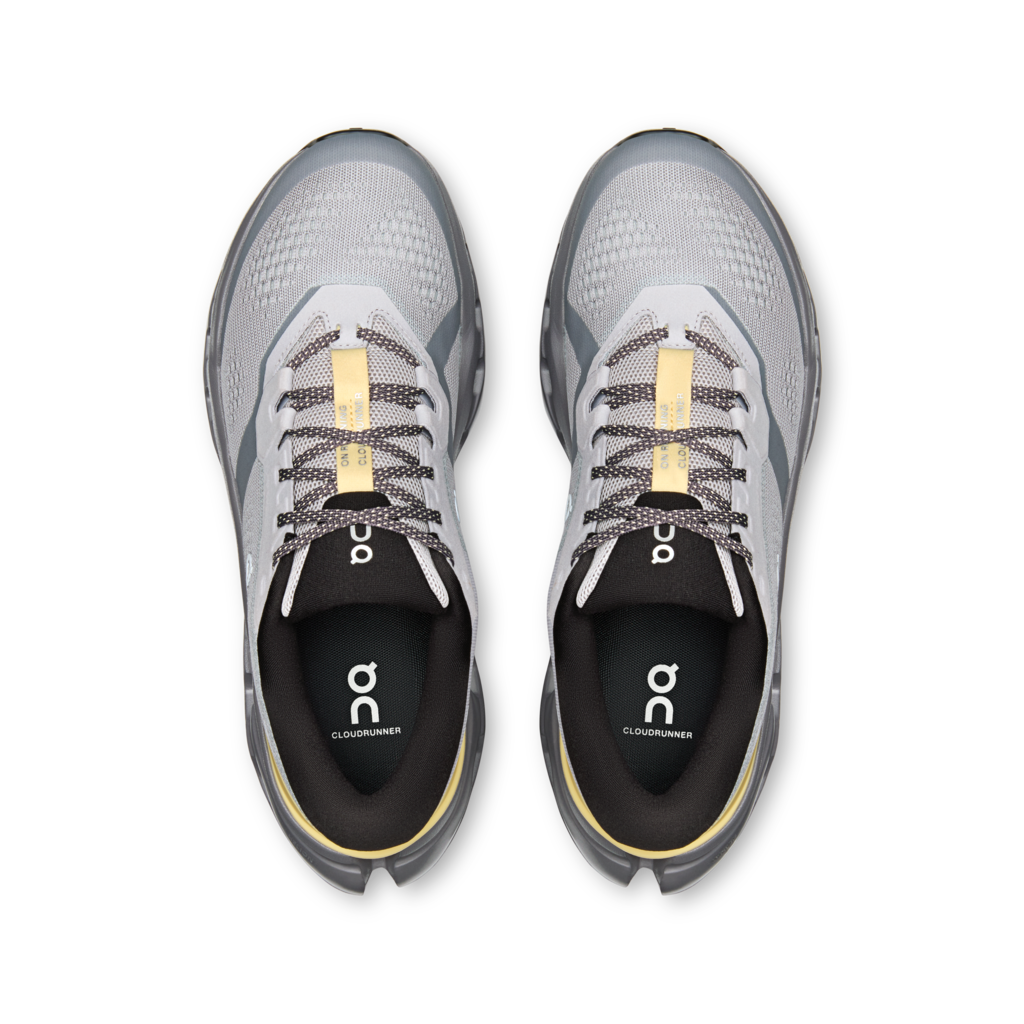 A pair of On Cloudrunner trail running shoes shown from a top-down view, featuring a light gray and white mesh upper with black and gray details, black laces, and a gold accent on the tongue and inner sole.