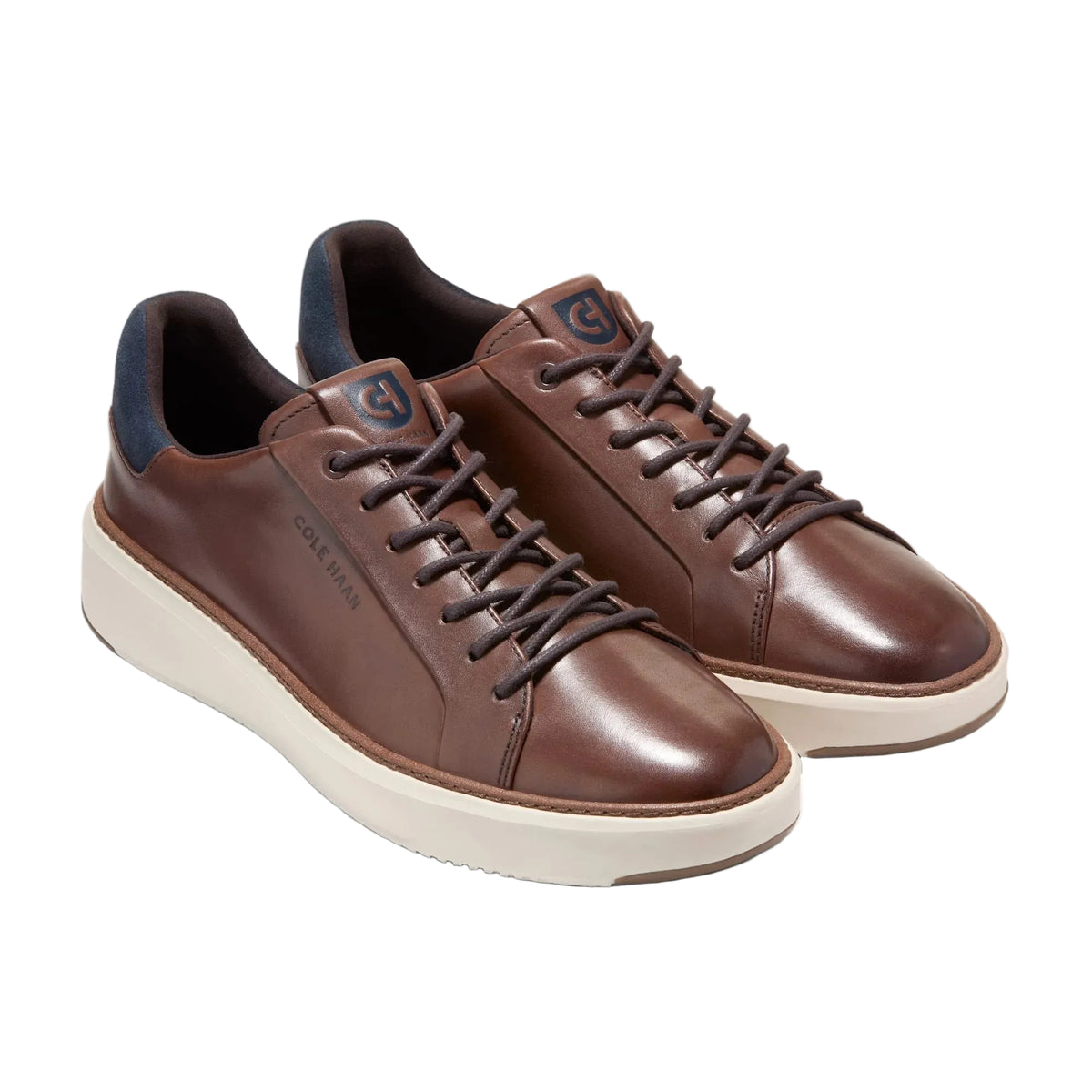A pair of brown leather low-top sneakers with dark brown laces, cream-colored soles, and dark navy blue lining, displayed on a light gray background.