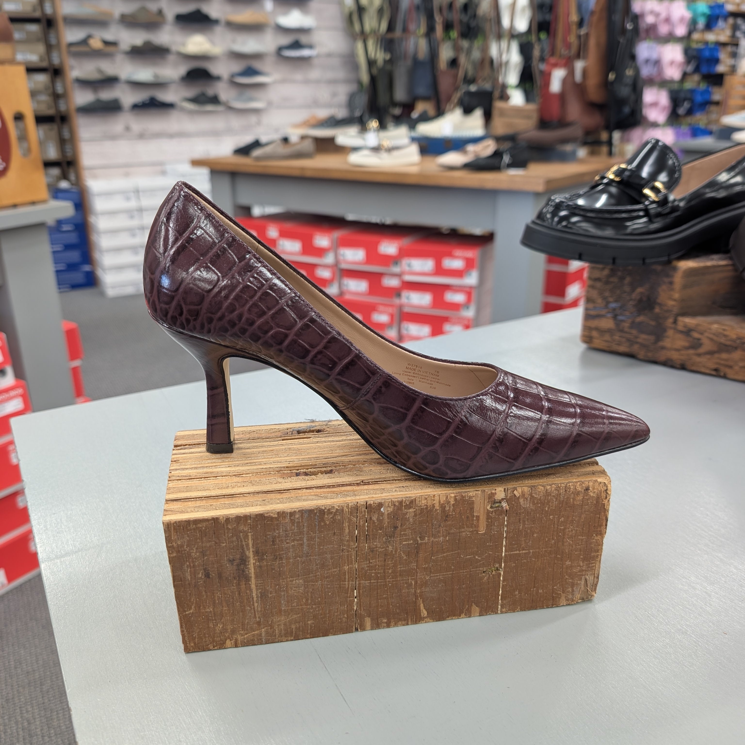 A dark burgundy crocodile-embossed pointed-toe pump with a mid-height stiletto heel, displayed on a wooden block in a shoe store.
