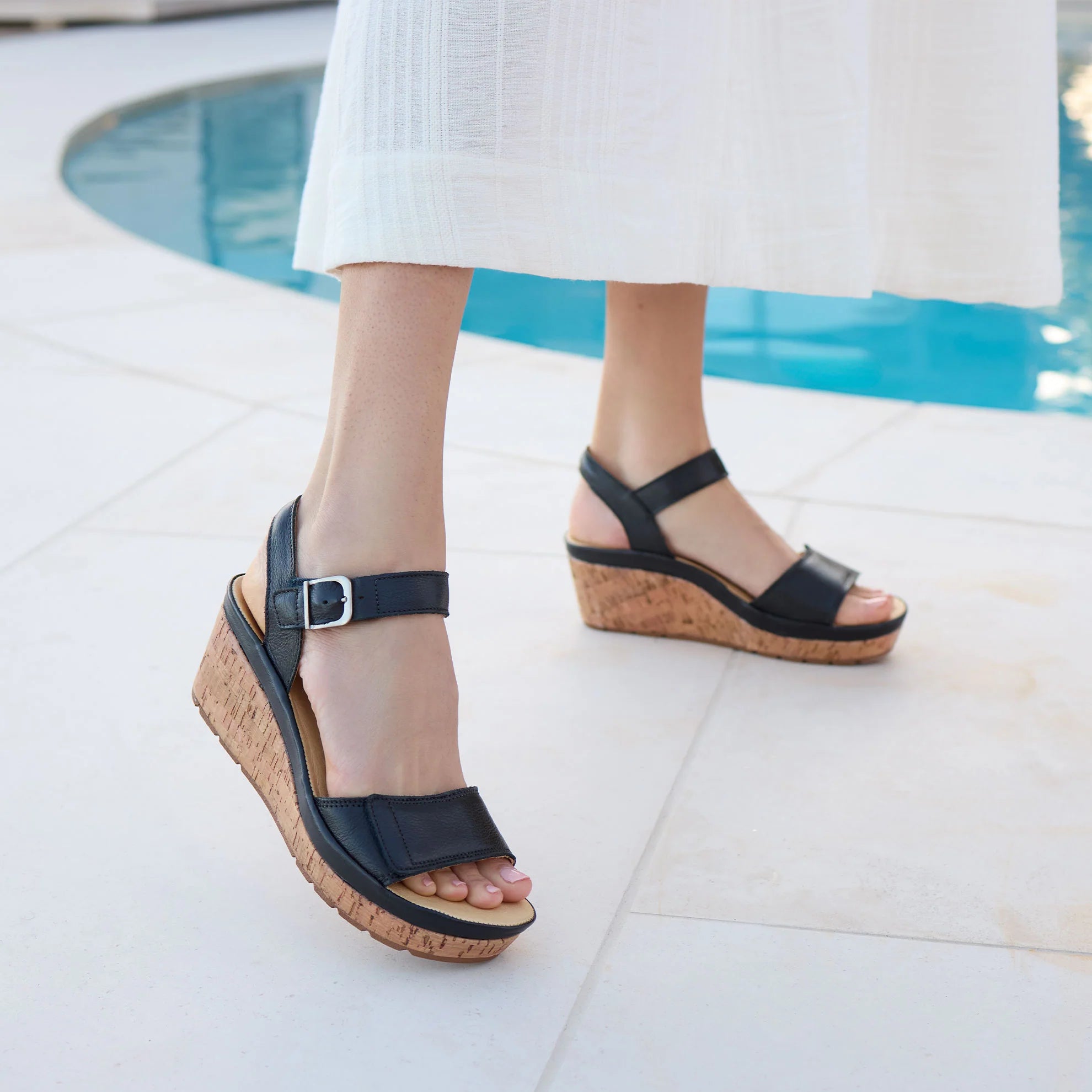 Black wedge sandals worn by a person standing on a tiled floor near a pool.