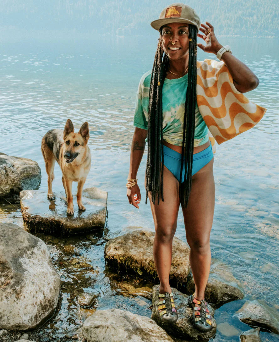 Woman in a swimsuit standing on rocks by a body of water with a dog.