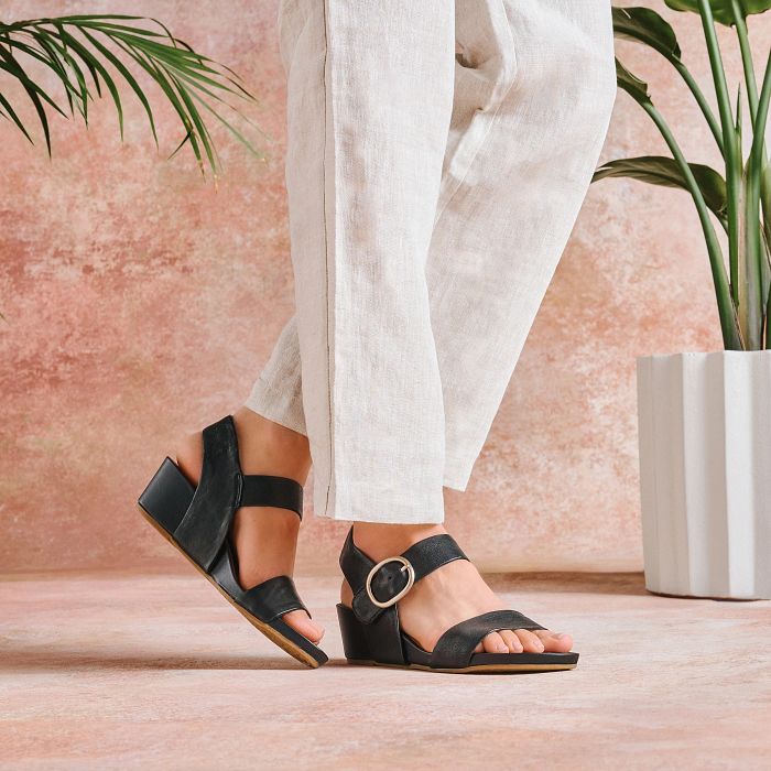 Black sandals worn with white pants against a pink wall with plants.