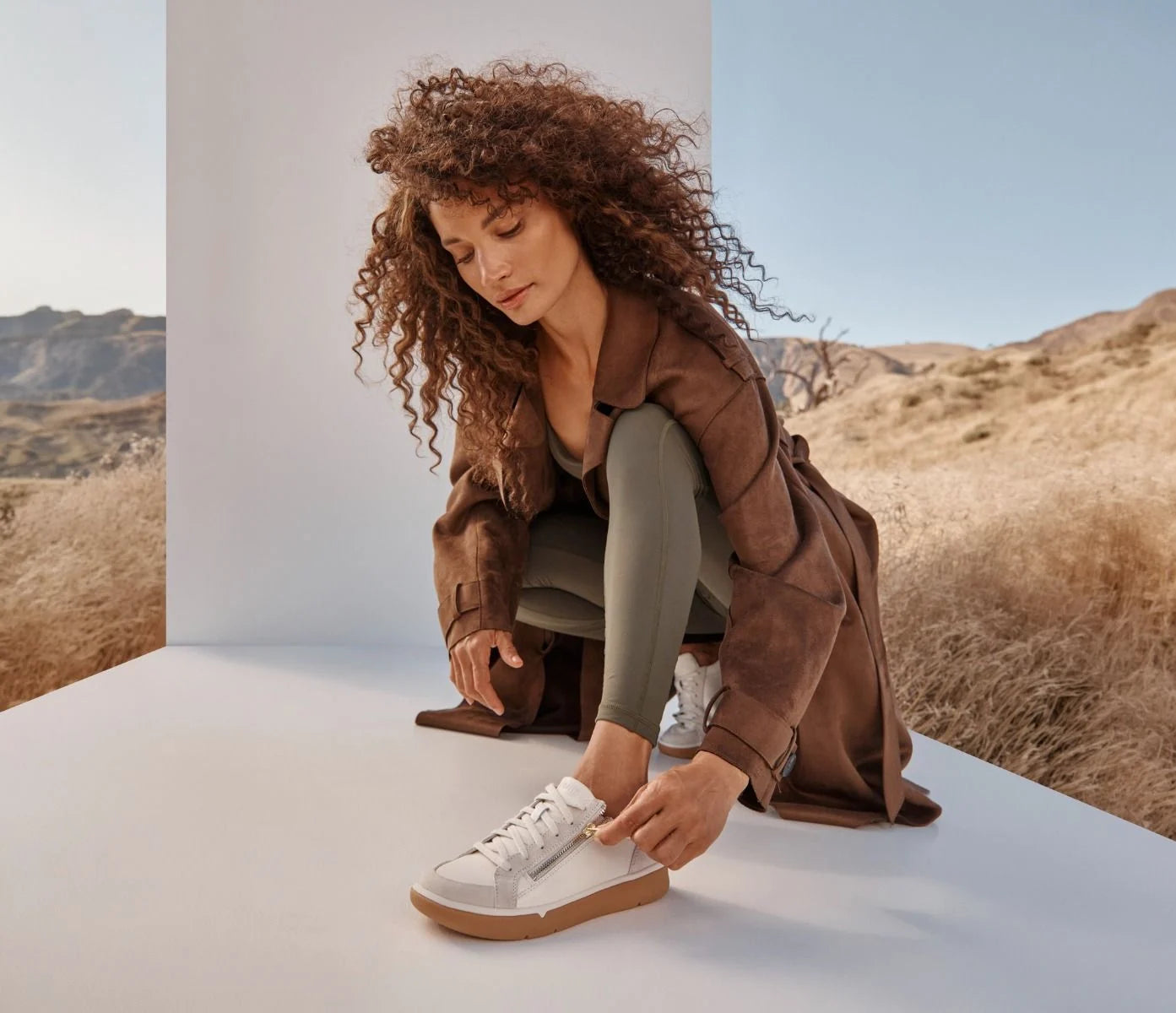 A woman with voluminous curly brown hair crouches on a white platform in a desert setting, wearing a brown jacket, olive green leggings, and white sneakers with gum soles.