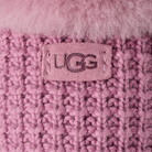 Close-up of a pink knit beanie with a fuzzy pink trim and a central rectangular UGG logo patch.
