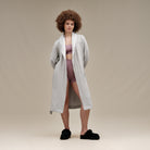 A woman stands against a plain beige studio background wearing a light gray open-front robe over a mauve-pink sports bra and matching high-waisted shorts, with black fuzzy slippers.