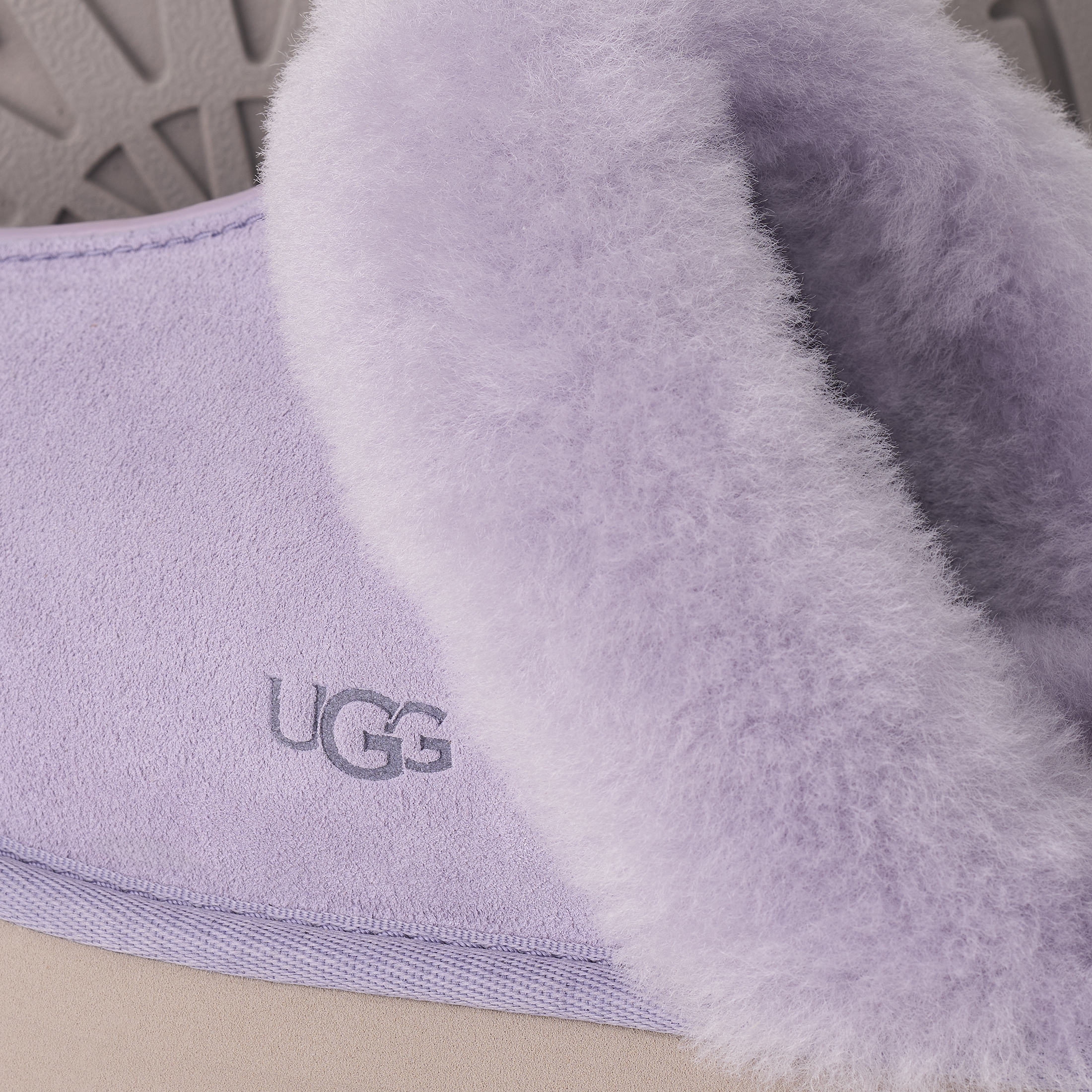 Close-up of a lavender UGG slipper showing the fuzzy shearling-style collar and the embroidered UGG logo on the suede side.