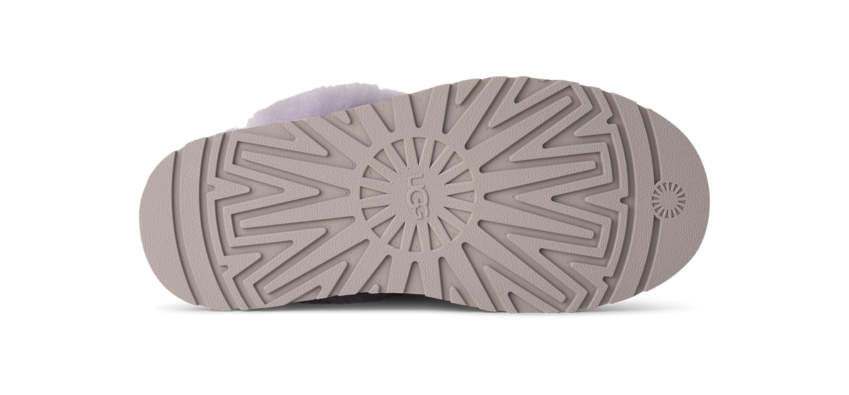 The sole of a UGG slipper in a taupe-brown color, showing the tread pattern and the embossed UGG logo in the center, with a light lavender fuzzy lining visible at the top edge.