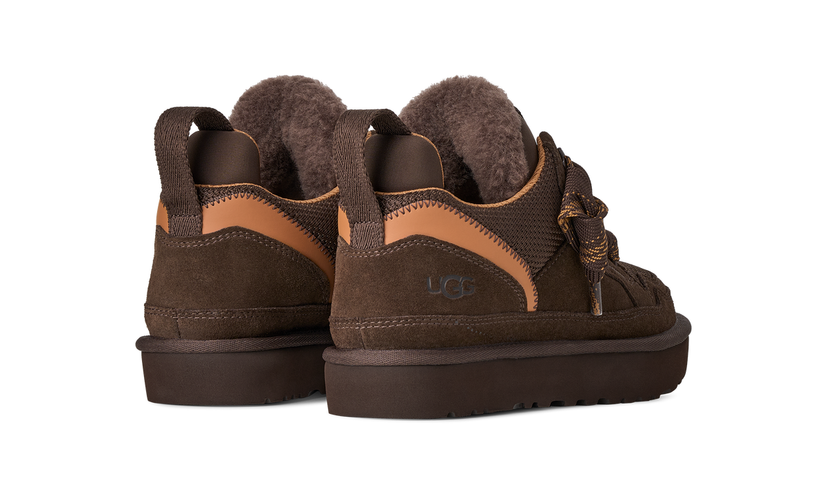 A pair of UGG low-top slip-on boots in dark brown suede and leather, featuring tan contrast panels and a plush brown faux-fur lining, shown from a three-quarters angle on a white background.