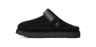 A single black suede clog-style slipper shown in side profile against a solid white background. The upper features a black suede body, a black sole, and a black strap with a textured geometric pattern and a small round button closure.