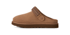A single brown suede clog-style slipper shown in side profile on a plain white background. The upper is a warm tan-brown suede, with a darker brown sole and a patterned brown strap detail across the back.