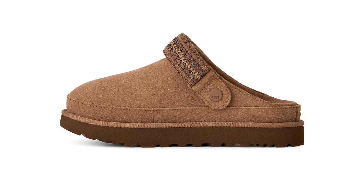 A single brown suede clog-style slipper shown in side profile on a plain white background. The upper is a warm tan-brown suede, with a darker brown sole and a patterned brown strap detail across the back.