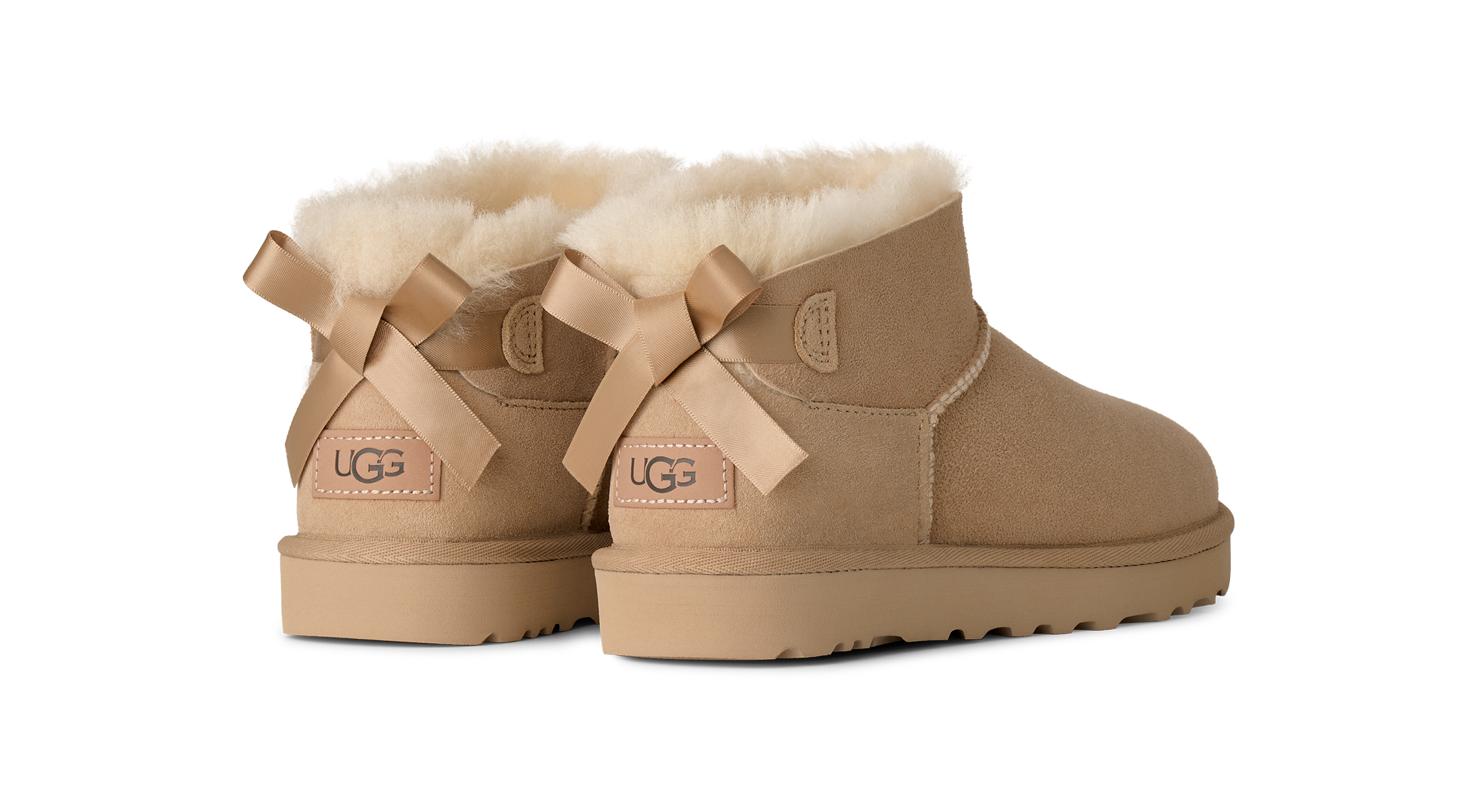 A pair of UGG Mini Bailey boots in tan suede with fluffy cream shearling lining and matching tan ribbon bows on the back, shown against a plain white background.