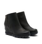 A pair of black leather wedge ankle boots with side zippers and chunky lug soles, shown on a light off-white background.