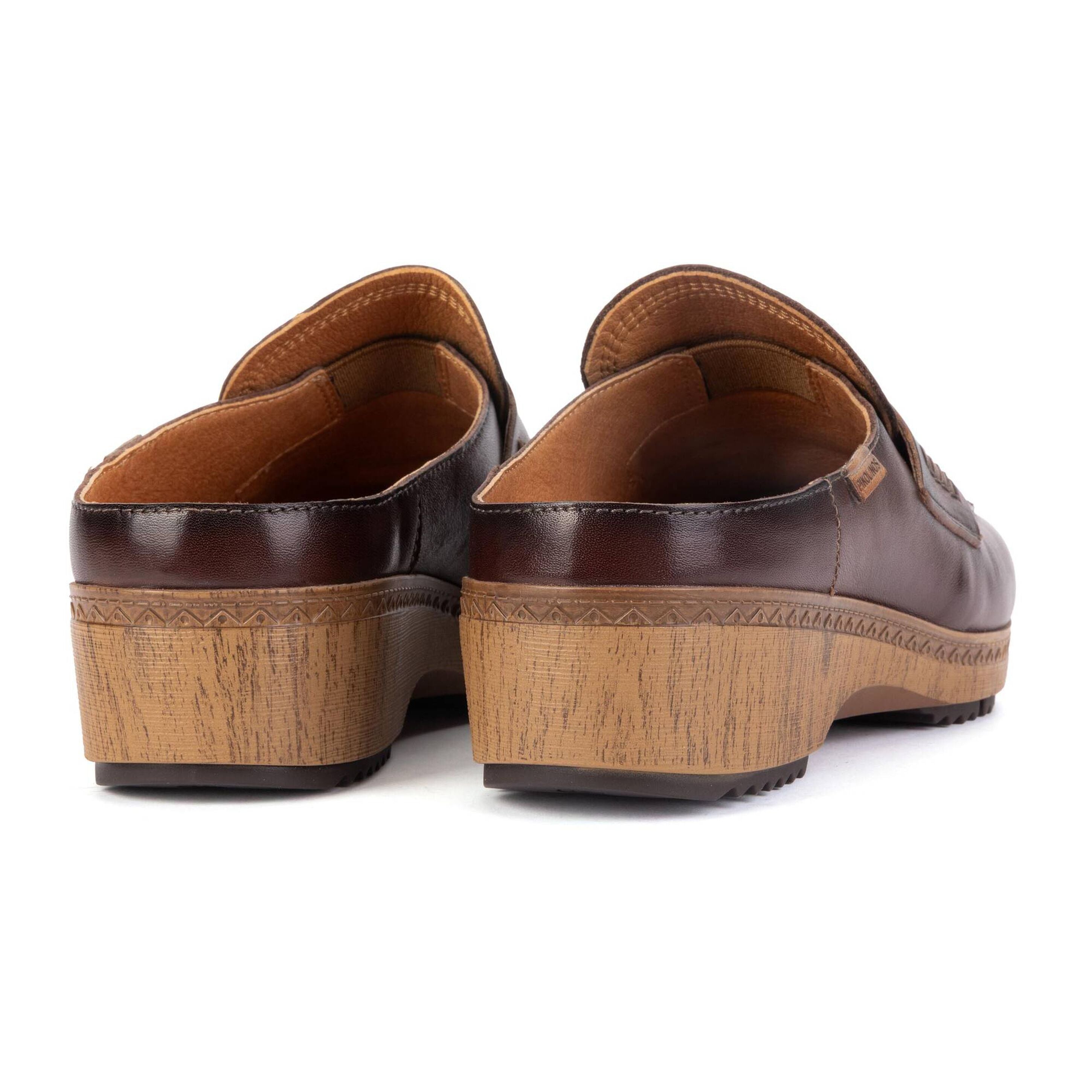 A pair of dark brown leather clogs with chunky, textured natural wood wedge soles, shown from a rear three-quarter angle on a white background.