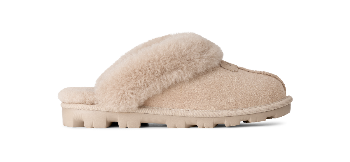 A single slip-on mule slipper in beige suede with a fluffy cream shearling-style strap and matching beige sole, shown in side profile on a white background.