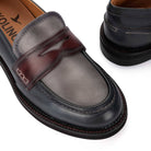 A close-up of a penny loafer in dark brown and black leather, showing the side profile with contrast stitching and a pull strap. A second shoe is partially visible in the background.