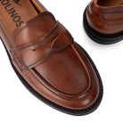 A close-up of a brown leather penny loafer shoe on a white background, with part of another matching shoe visible in the upper right. The insole shows the brand name OLIVOS.
