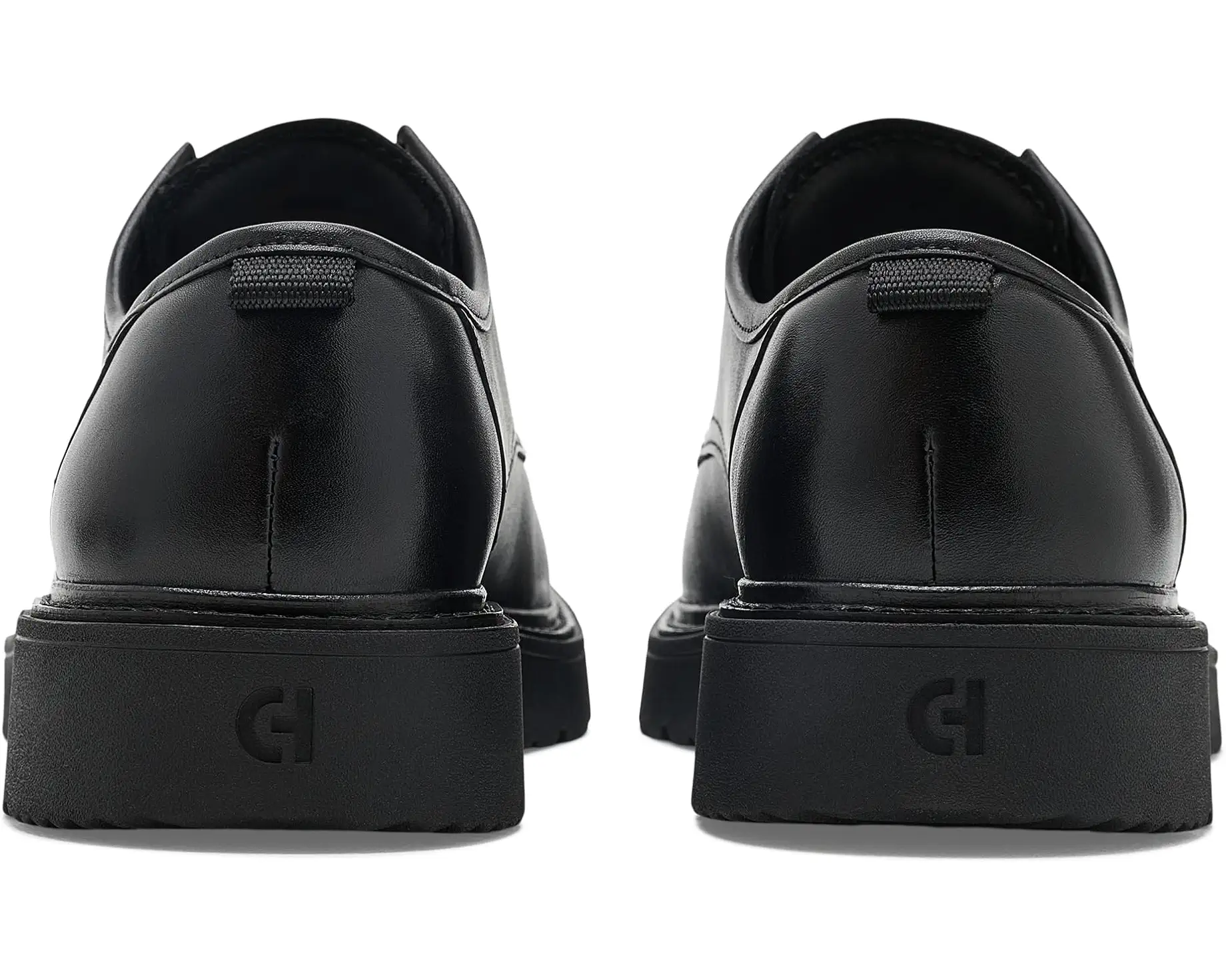 A pair of black leather lace-up shoes shown from the back, featuring thick black platform soles with a debossed monogram logo, photographed on a clean white background.