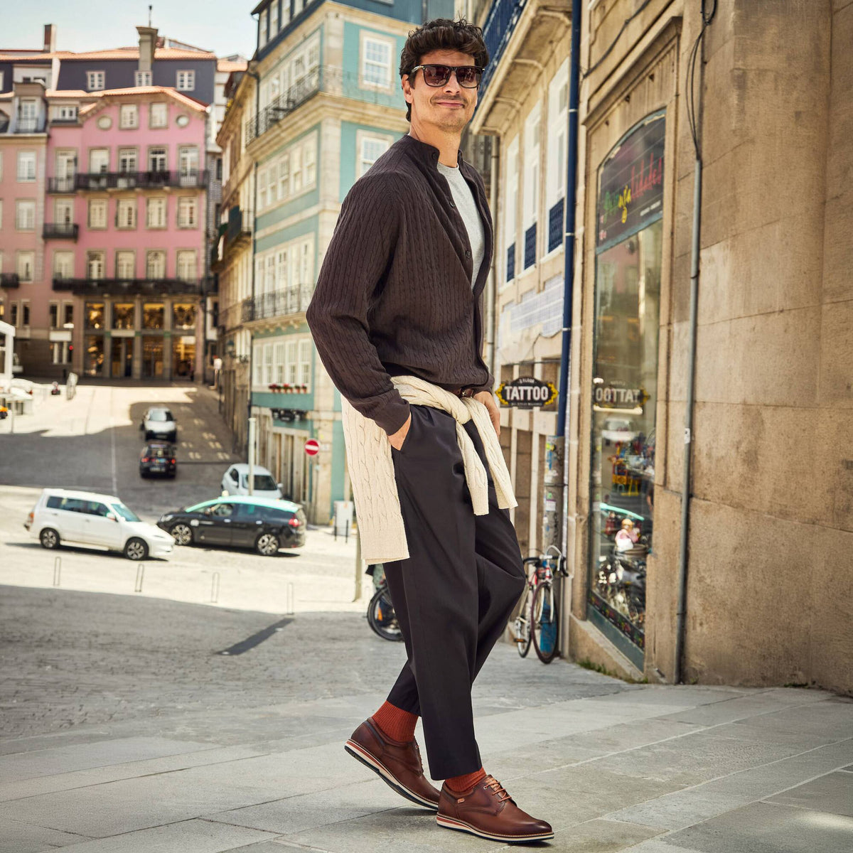 A man wearing a dark brown ribbed cardigan, dark trousers, brown leather shoes, and red socks walks down a cobblestone street in front of colorful European buildings.
