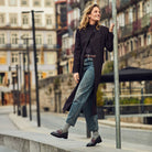 A woman stands on a city sidewalk leaning against a metal pole, wearing a long black coat, blue jeans, gray socks, and black loafers. The background shows blurred urban buildings and street elements.