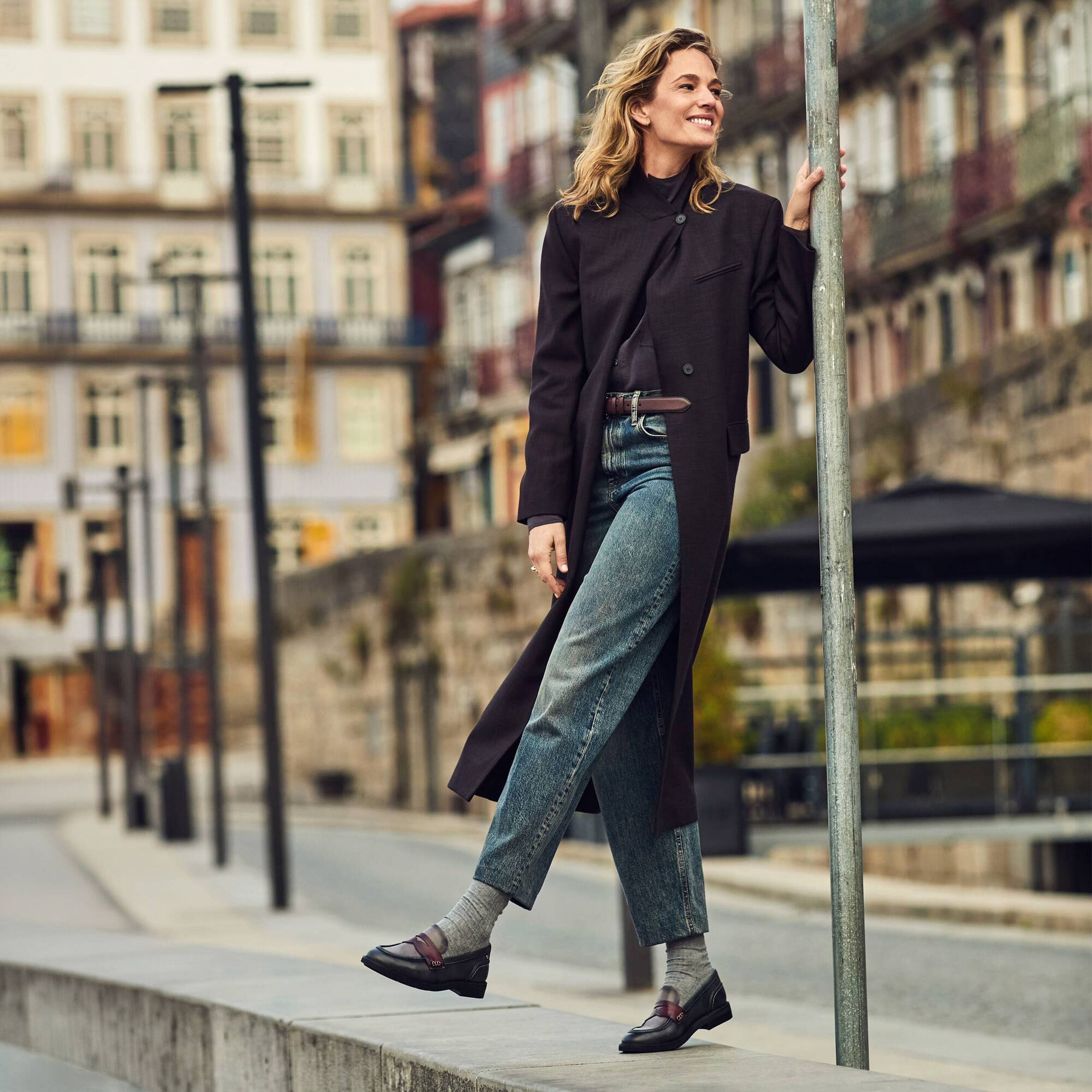 A woman stands on a city sidewalk leaning against a metal pole, wearing a long black coat, blue jeans, gray socks, and black loafers. The background shows blurred urban buildings and street elements.