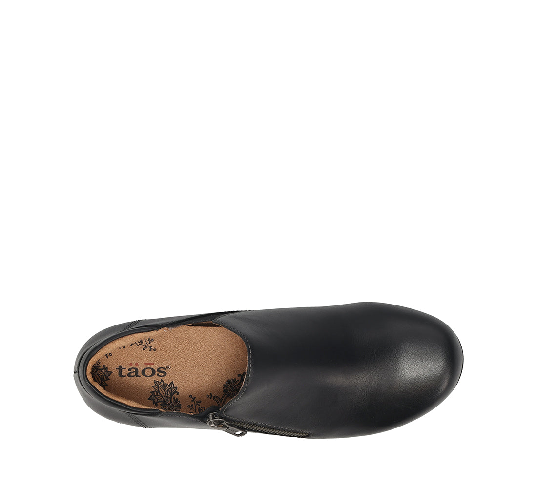 A black slip-on shoe shown from a top-down view on a plain white background, with a visible tan insole printed with the brand name 'taos'.
