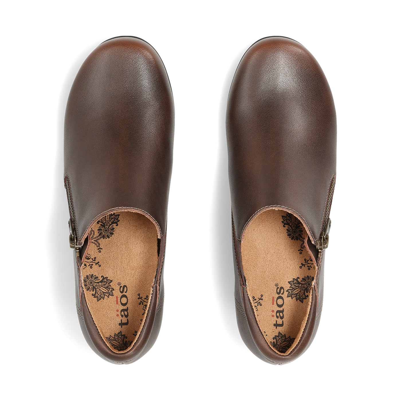 A pair of dark brown leather slip-on shoes shown from a top-down view on a white background. The brand name 'taos' is printed on the tan insoles with a black floral pattern.