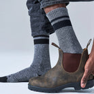 A person wearing gray heathered crew socks with black stripes and a brown Chelsea-style boot, with one hand holding the boot open. The background is a clean light gray studio backdrop.