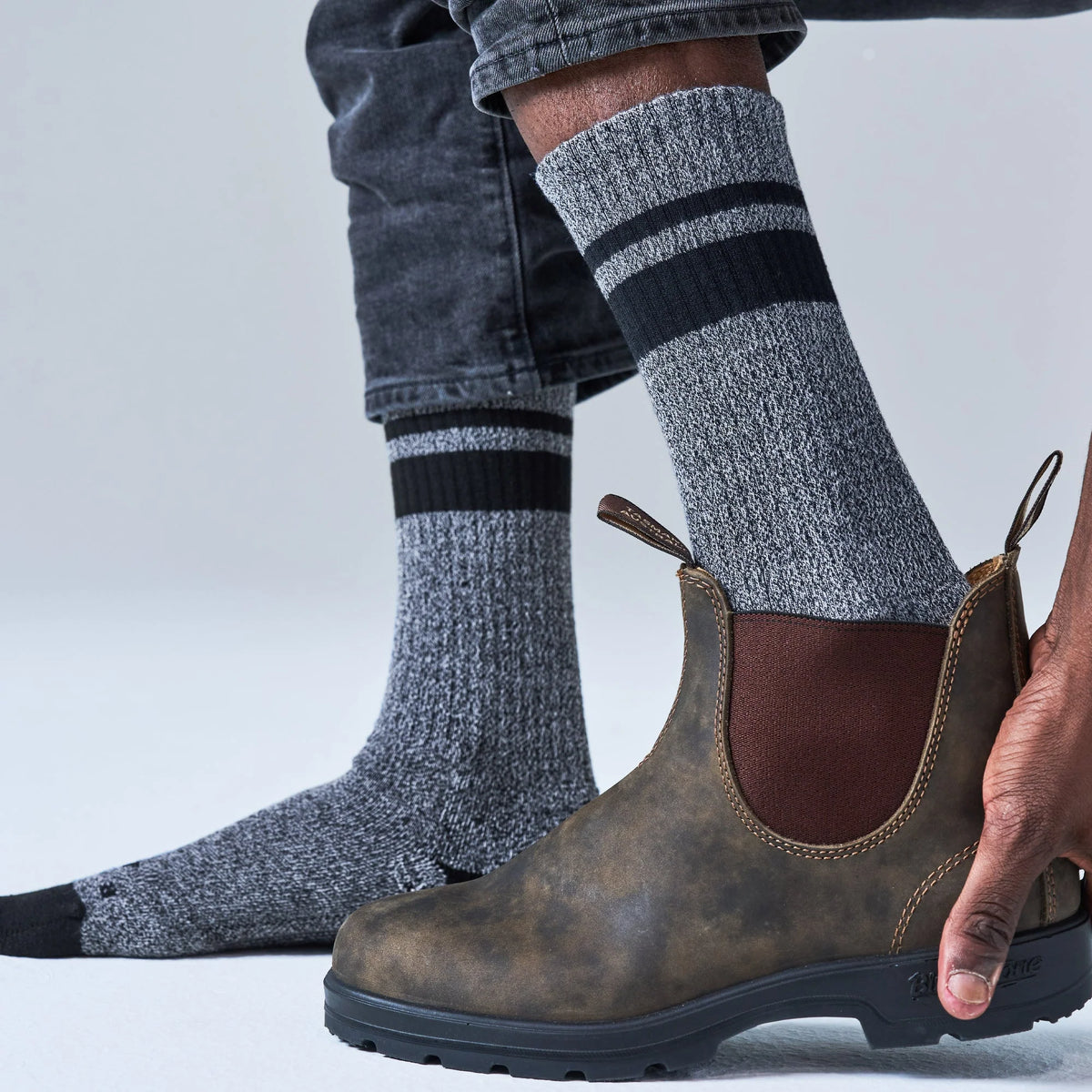A person wearing gray heathered crew socks with black stripes and a brown Chelsea-style boot, with one hand holding the boot open. The background is a clean light gray studio backdrop.