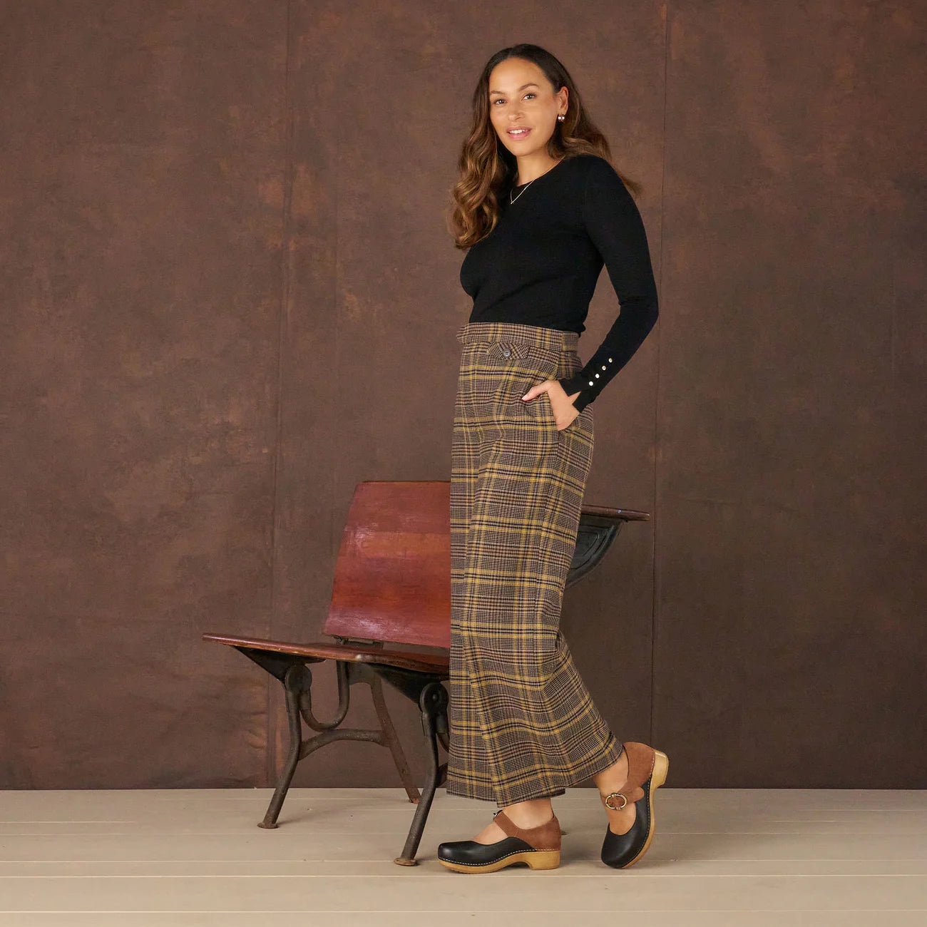 Woman wearing a black top and plaid pants standing against a brown textured wall.
