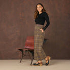 Woman wearing a black top and plaid pants standing against a brown textured wall.