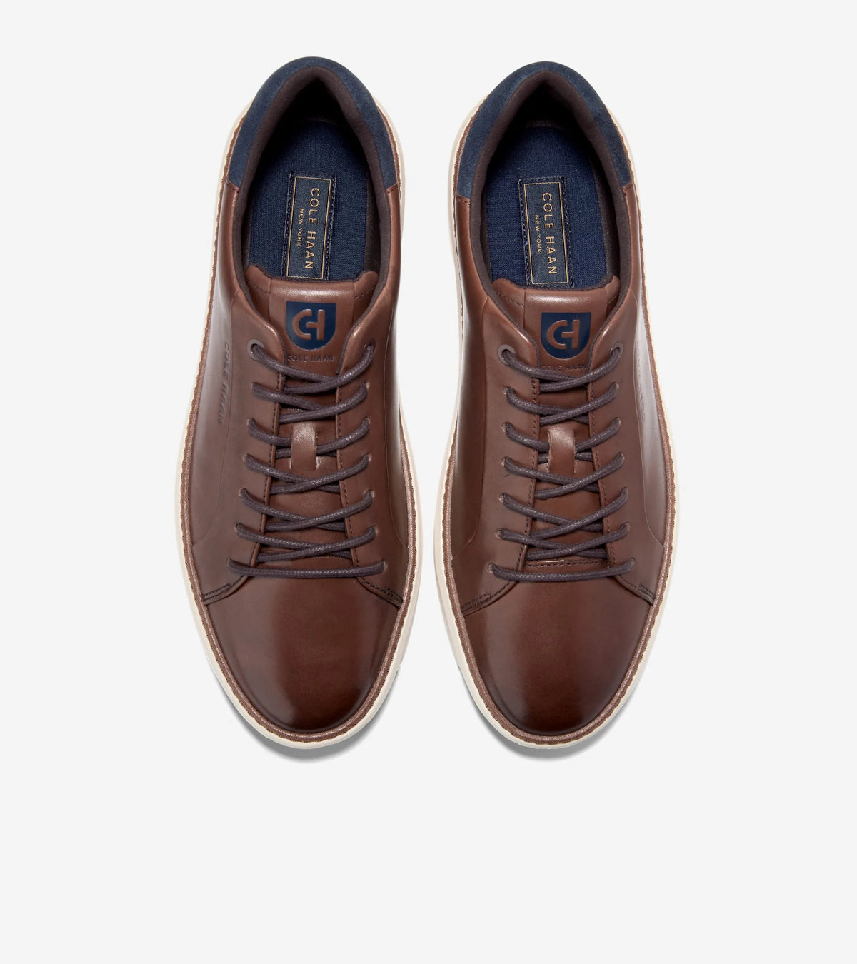 A pair of brown leather low-top sneakers shown from a top-down view on a light gray background. The shoes feature dark brown laces, a dark blue interior lining, and a white midsole.
