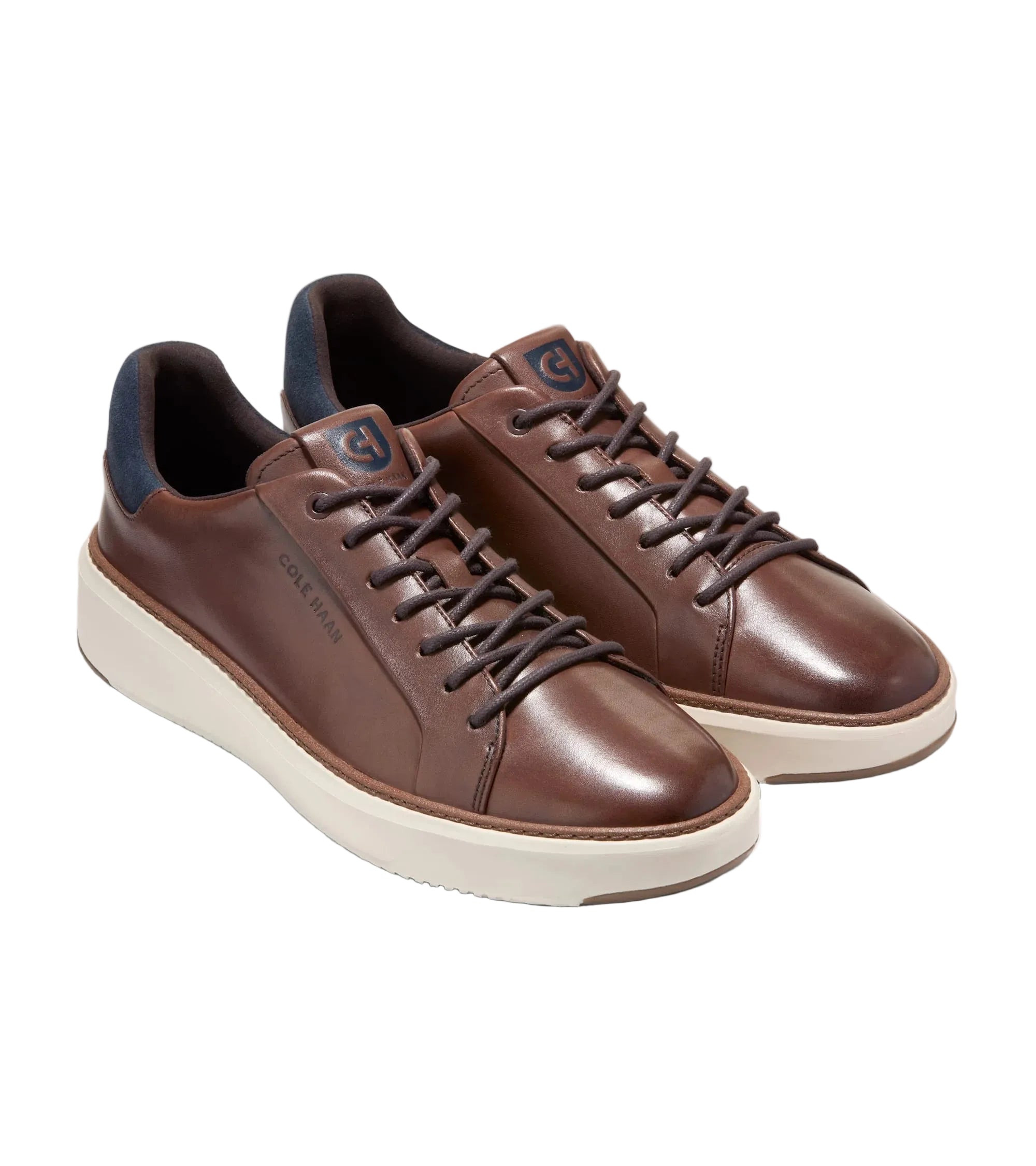 A pair of brown leather low-top sneakers with dark brown laces, cream-colored soles, and dark navy blue lining, displayed on a light gray background.