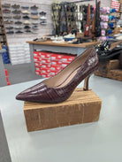 A single pointed-toe high heel pump in a dark burgundy crocodile-embossed leather, displayed on a wooden block inside a shoe store.