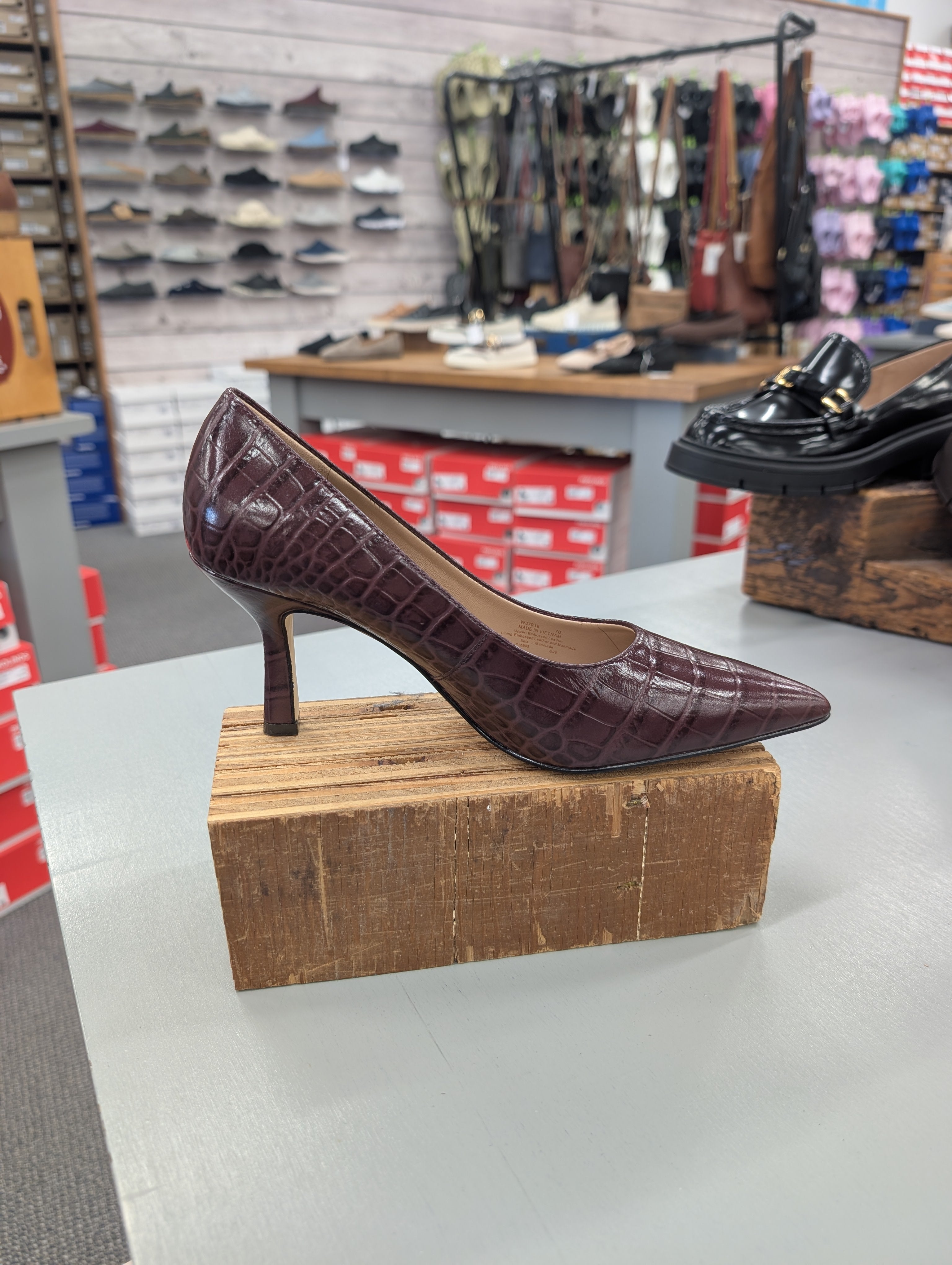 A dark burgundy crocodile-embossed pointed-toe pump with a mid-height stiletto heel, displayed on a wooden block in a shoe store.
