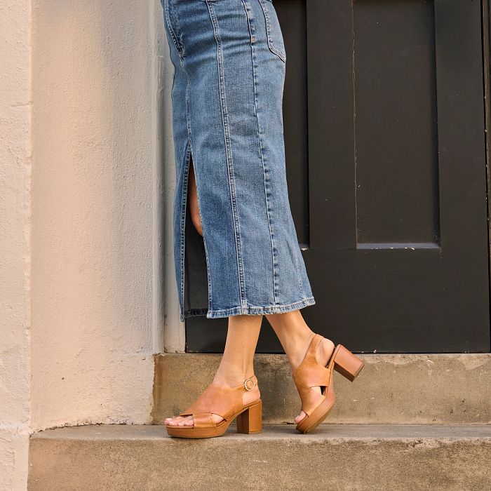 Person wearing blue jeans and brown sandals standing against a building.