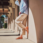 Person wearing a light blue shirt, beige pants, and brown shoes leaning against a wall in an outdoor setting.