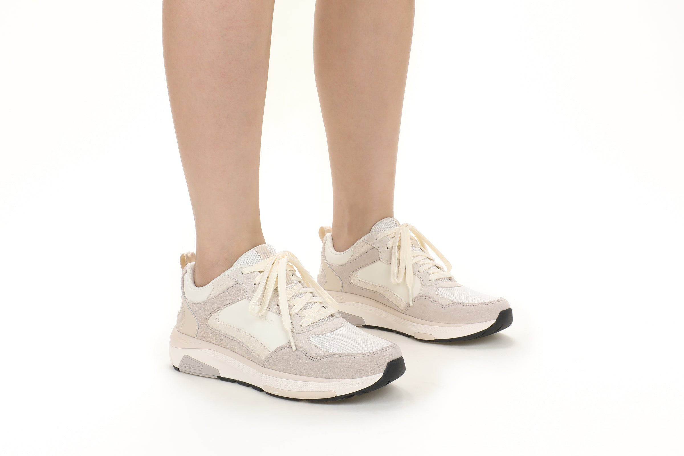 A pair of low-top athletic sneakers in off-white, beige, and light gray, shown on a model's feet against a clean white background.
